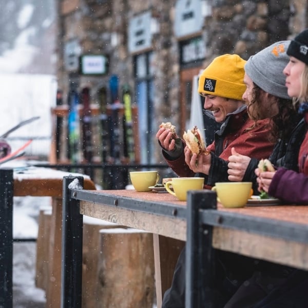 Three people sit on outdoor patio eating and drinking in the winter. It is snowing lightly and they are dressed in winter clothes.