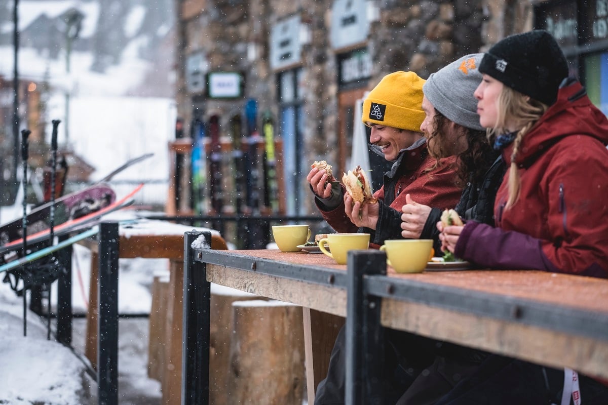 Three people sit on outdoor patio eating and drinking in the winter. It is snowing lightly and they are dressed in winter clothes.