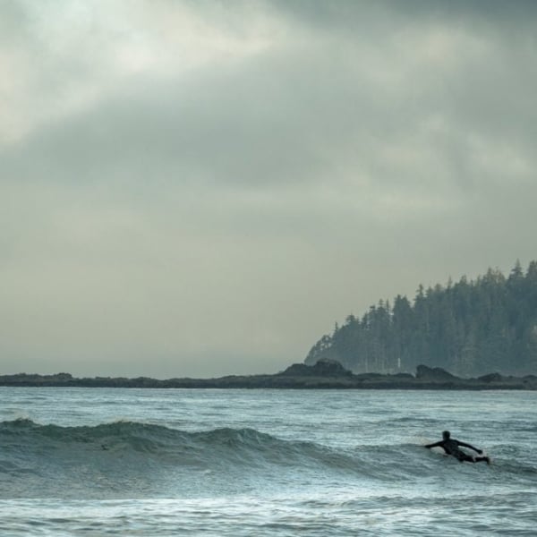 A surfer paddling out over the waves in overcast weather. There is an island in the far distance on the right and a few clouds in the sky.