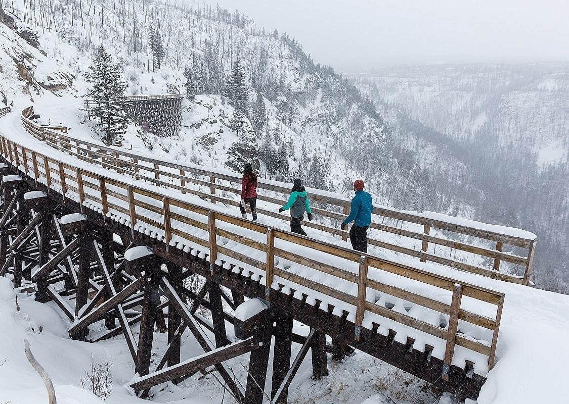 Tres personas con raquetas de nieve caminan por un puente de madera cubierto de nieve en el Kettle Valley Rail Trail