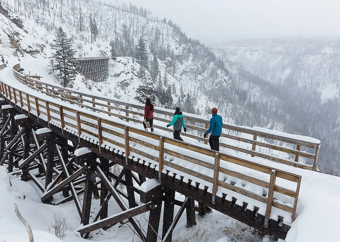 Drei Schneeschuhwander*innen gehen über eine schneebedeckte Holzbrücke auf dem Kettle Valley Rail Trail.