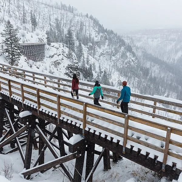 Drei Schneeschuhwander*innen gehen über eine schneebedeckte Holzbrücke auf dem Kettle Valley Rail Trail.