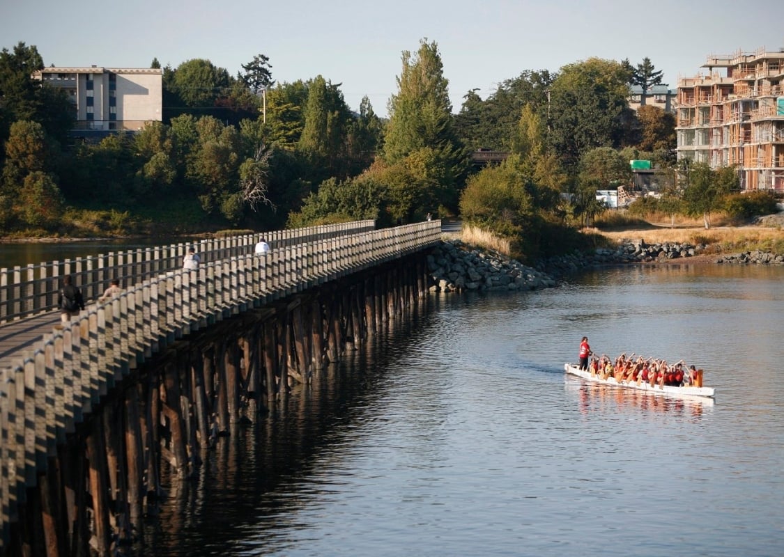 A dragon boat paddles in the water away from a bridge. Cyclists are seen riding over the bridge on a clear, sunny day.