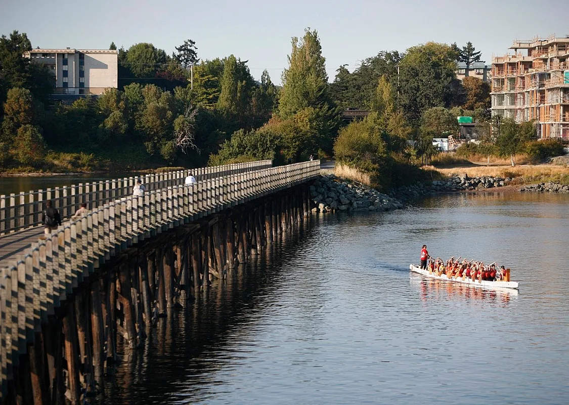A dragon boat paddles in the water away from a bridge. Cyclists are seen riding over the bridge on a clear, sunny day.