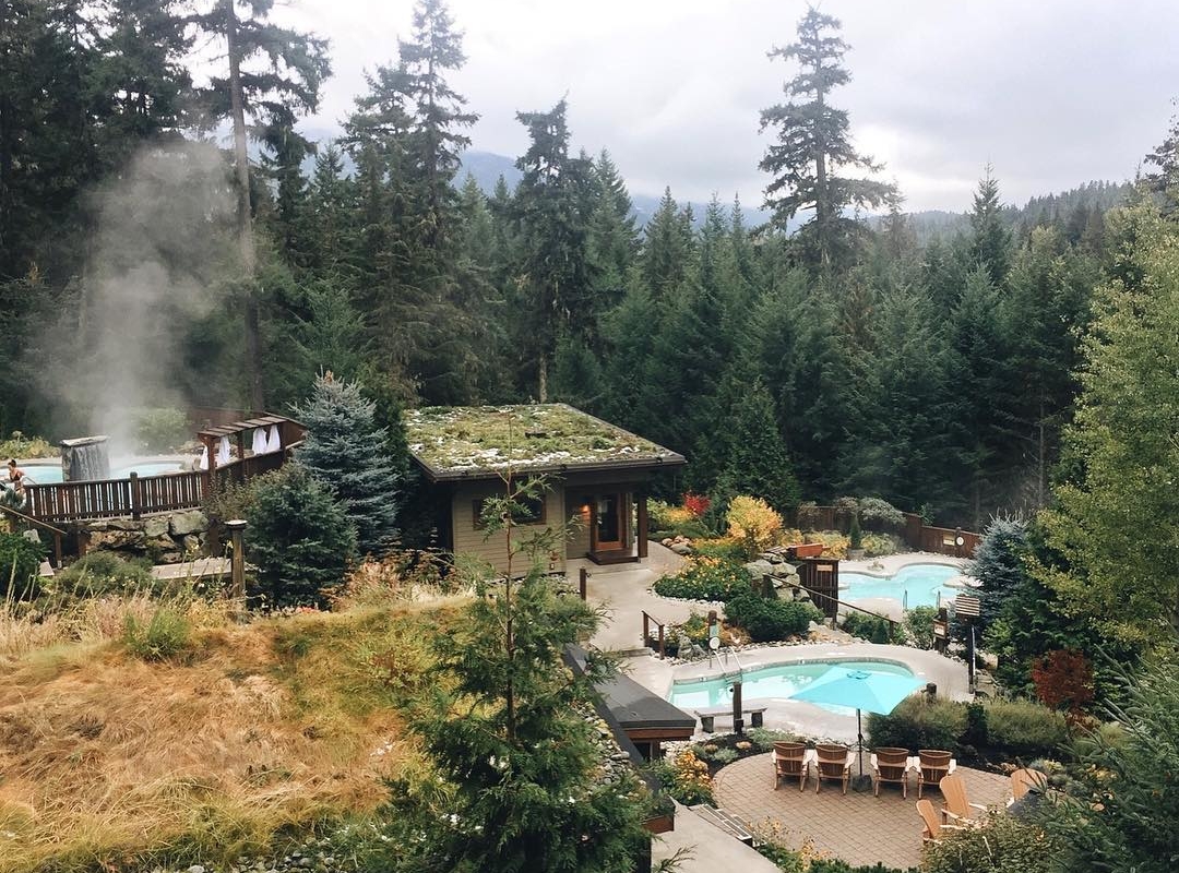 Overview of Scandinave Spa in Whistler on an overcast day. There are two pools on the property, lots of trees and flowers surrounding the pools and a building that contains the sauna and steam room.