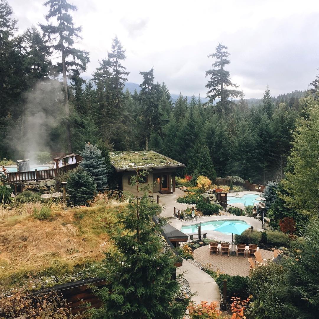 Overview of Scandinave Spa in Whistler on an overcast day. There are two pools on the property, lots of trees and flowers surrounding the pools and a building that contains the sauna and steam room.