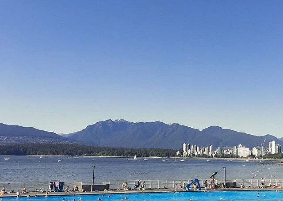 Twenty or so people swim in an aqua-coloured pool overlooking the darker blue of the ocean. The mountains and downtown Vancouver can be seen across the bay.