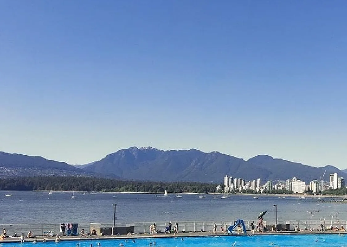 Twenty or so people swim in an aqua-coloured pool overlooking the darker blue of the ocean. The mountains and downtown Vancouver can be seen across the bay.