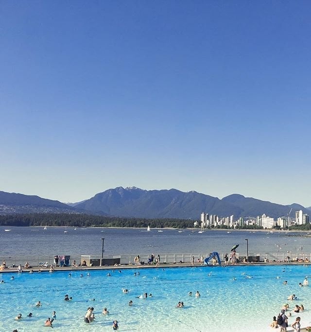 Twenty or so people swim in an aqua-coloured pool overlooking the darker blue of the ocean. The mountains and downtown Vancouver can be seen across the bay.