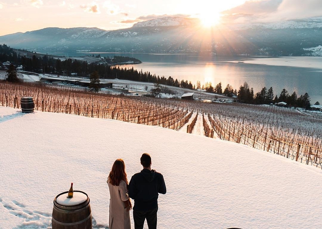 Un hombre y una mujer están juntos en la nieve, mirando hacia el viñedo y el lago en 50 Parallel Estate Winery