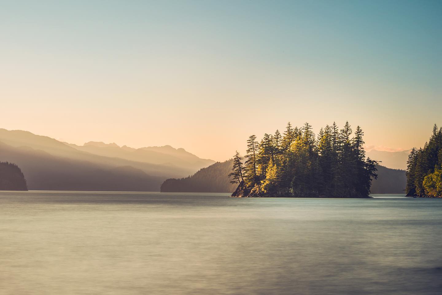 View over Harrison Lake with small islands nearby and mountains in the distance. The sun has disappeared and there's a pink-orange glow on the horizon