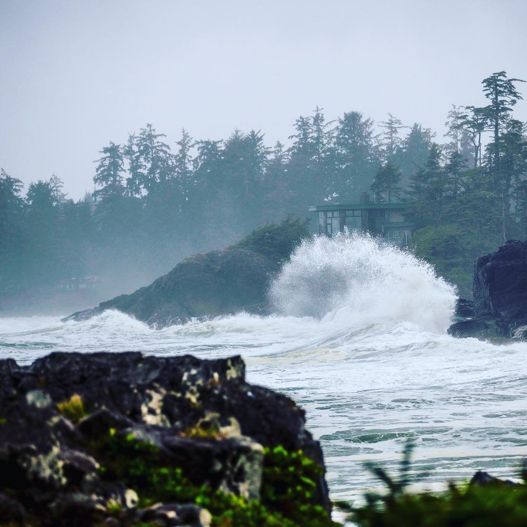 A wave crashes on the rocky shoreline with trees in the background on a stormy day in Tofino