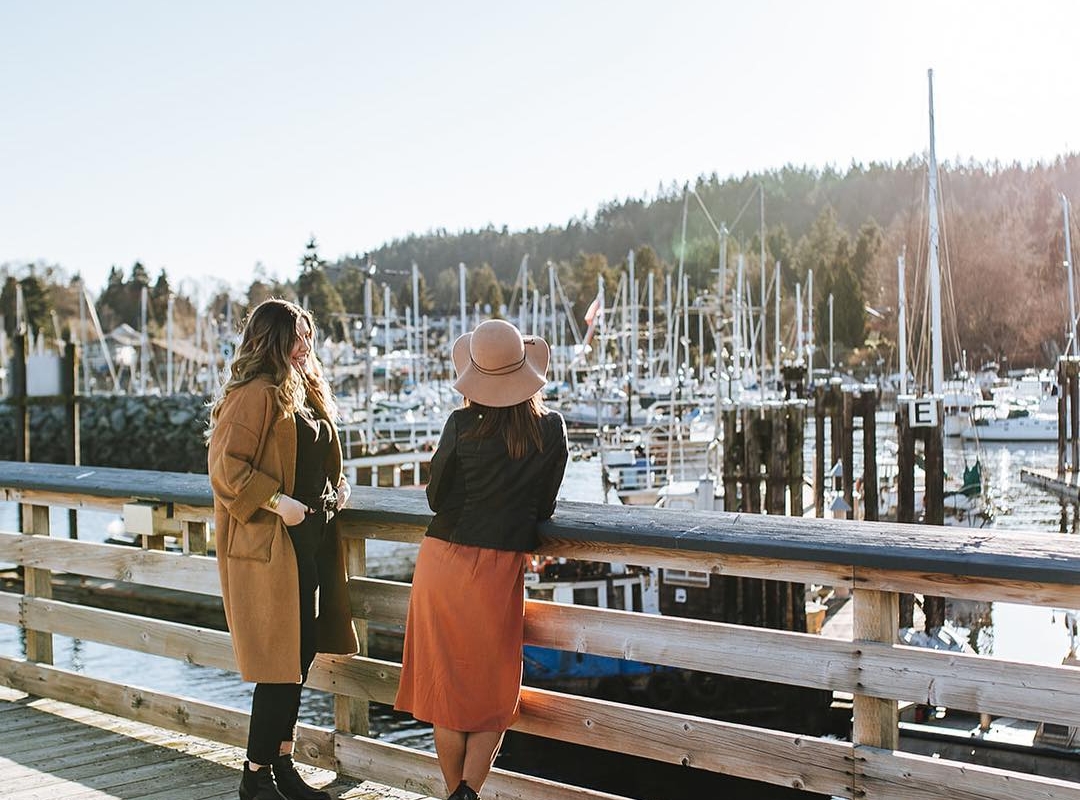 Two people stand on a pier looking out at the boats in the harbour at Gibsons Marina on a sunny day.