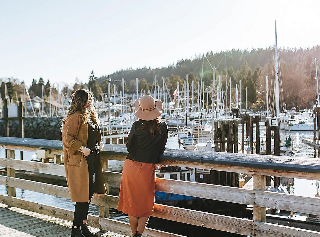 Two people stand on a pier looking out at the boats in the harbour at Gibsons Marina on a sunny day.