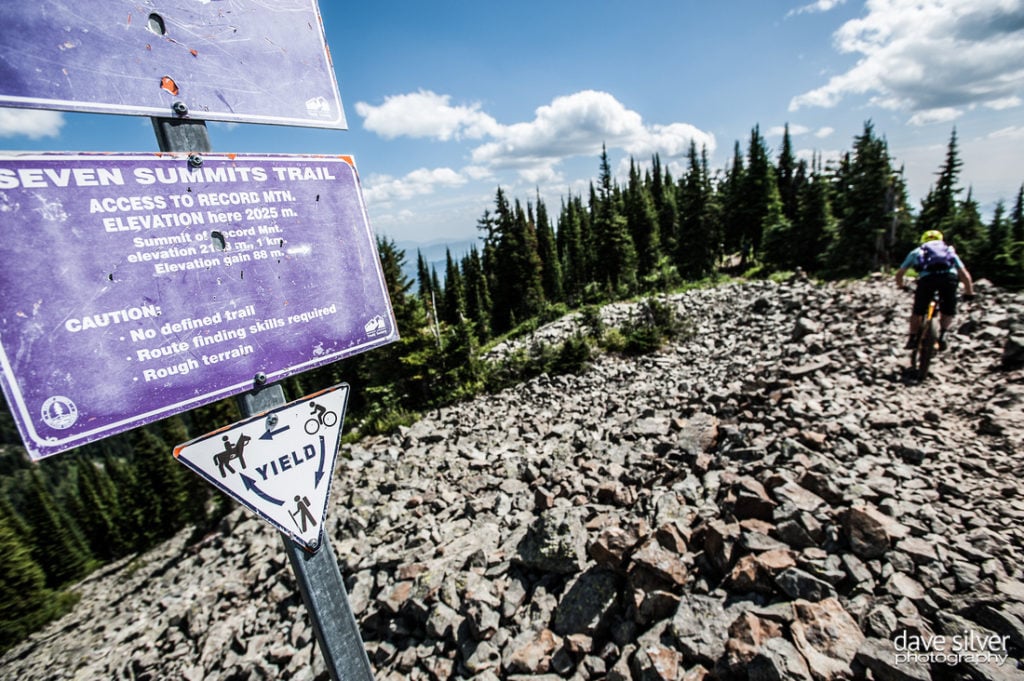 A mountain biker rides uphill over large rocks, past a blue sign that indicates Seven Summits Trail. The treeline is visible in front of them.
