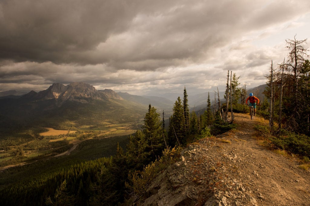 Mountain biker rides along a high mountain ridge with a green valley below and mountains in the background. The cloudy sky has a 
