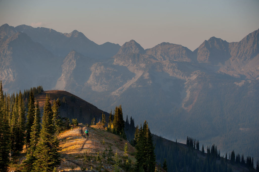 A person rides a mountain bike along the crest of a mountain with huge peaks in the background.