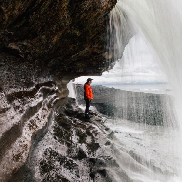 Una persona con una chaqueta roja de pie sobre una superficie rocosa y curva bajo el flujo de una cascada que cae desde arriba