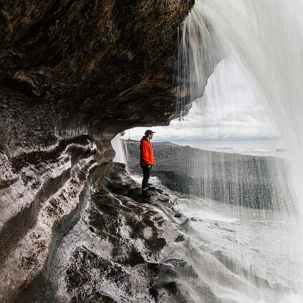 Person in a red jacket standing on a curved, rocky surface under the flow of a waterfall that cascades from above