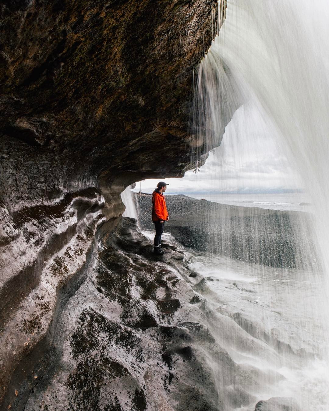Person in a red jacket standing on a curved, rocky surface under the flow of a waterfall that cascades from above