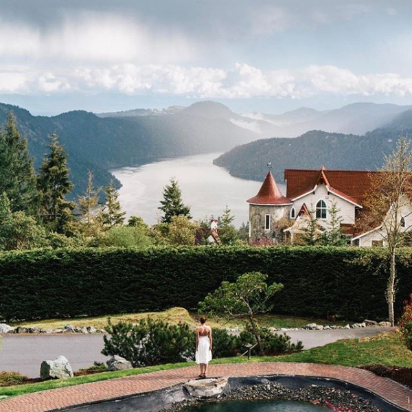 A woman stands on a red brick path taking in the views over the lake and mountains from Villa Eyrie Resort. There is a small pond behind here. The resort has a red roof and white walls.