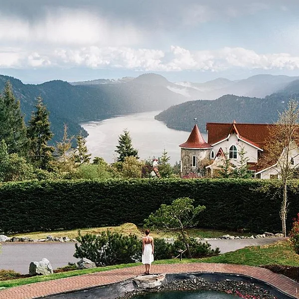 A woman stands on a red brick path taking in the views over the lake and mountains from Villa Eyrie Resort. There is a small pond behind here. The resort has a red roof and white walls.