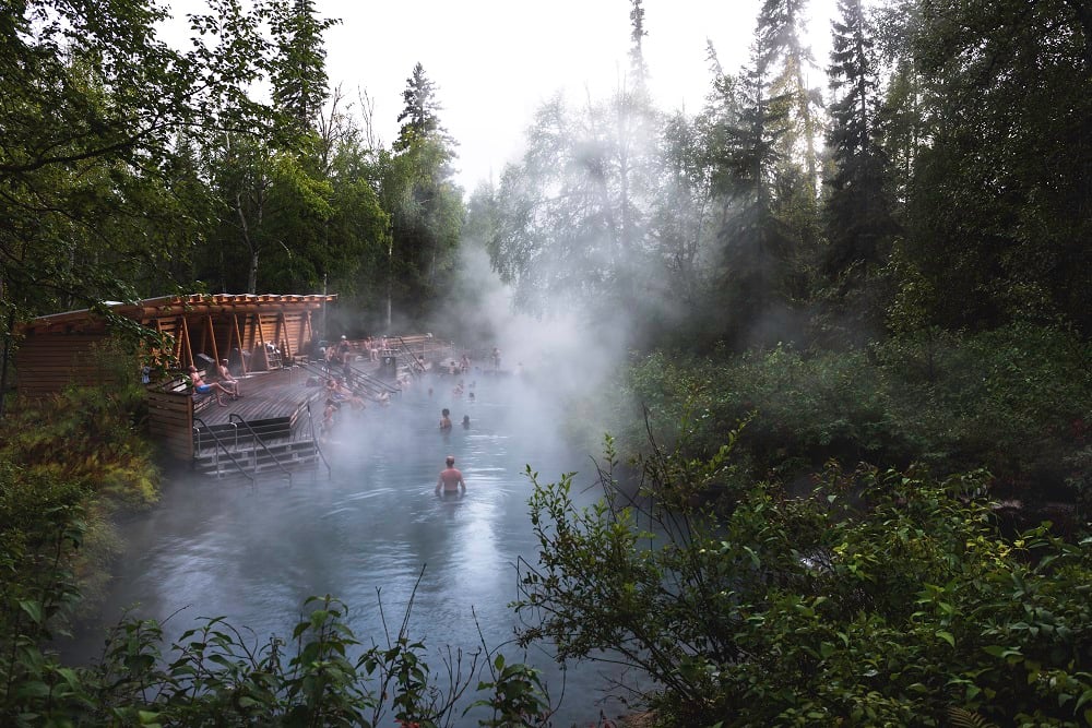 A steaming natural hot springs pool is surrounded by forest with a wooden deck and a wooden building on the left. Twenty or so people are in the water, with another five or six on the deck.
