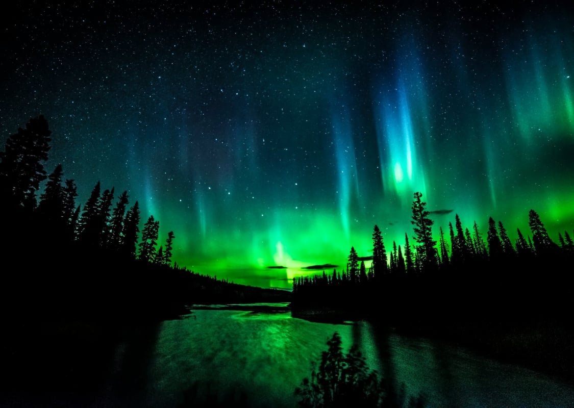 The vibrant green Northern Lights sparkles above a silhouette of a river and forest.