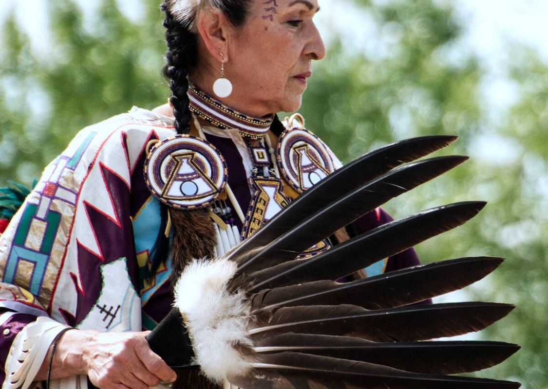 A dancer from Leona Smith - Muskwa River Fancy Dancers - Fort Nelson First Nation, dressed in vibrant traditional attire holds a feather fan.