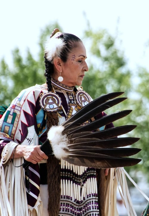 A dancer from Leona Smith - Muskwa River Fancy Dancers - Fort Nelson First Nation, dressed in vibrant traditional attire holds a feather fan.