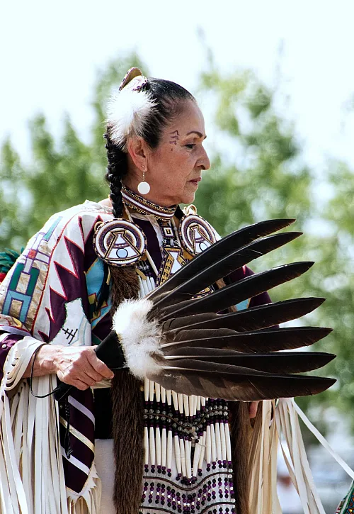 A dancer from Leona Smith - Muskwa River Fancy Dancers - Fort Nelson First Nation, dressed in vibrant traditional attire holds a feather fan.