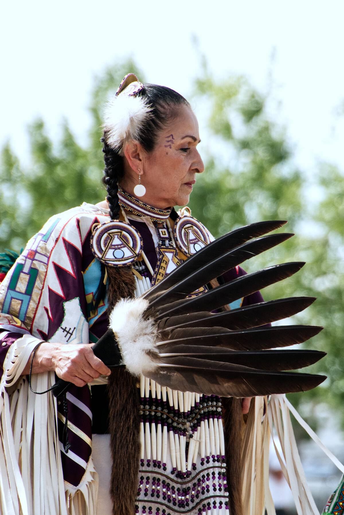A dancer from Leona Smith - Muskwa River Fancy Dancers - Fort Nelson First Nation, dressed in vibrant traditional attire holds a feather fan.