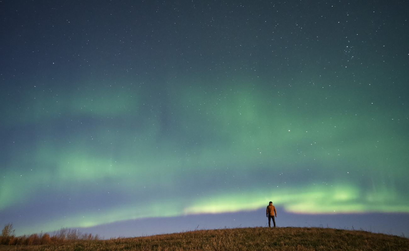 A person stands on a vast expanse of land looking up as the Northern Lights illuminate the sky.