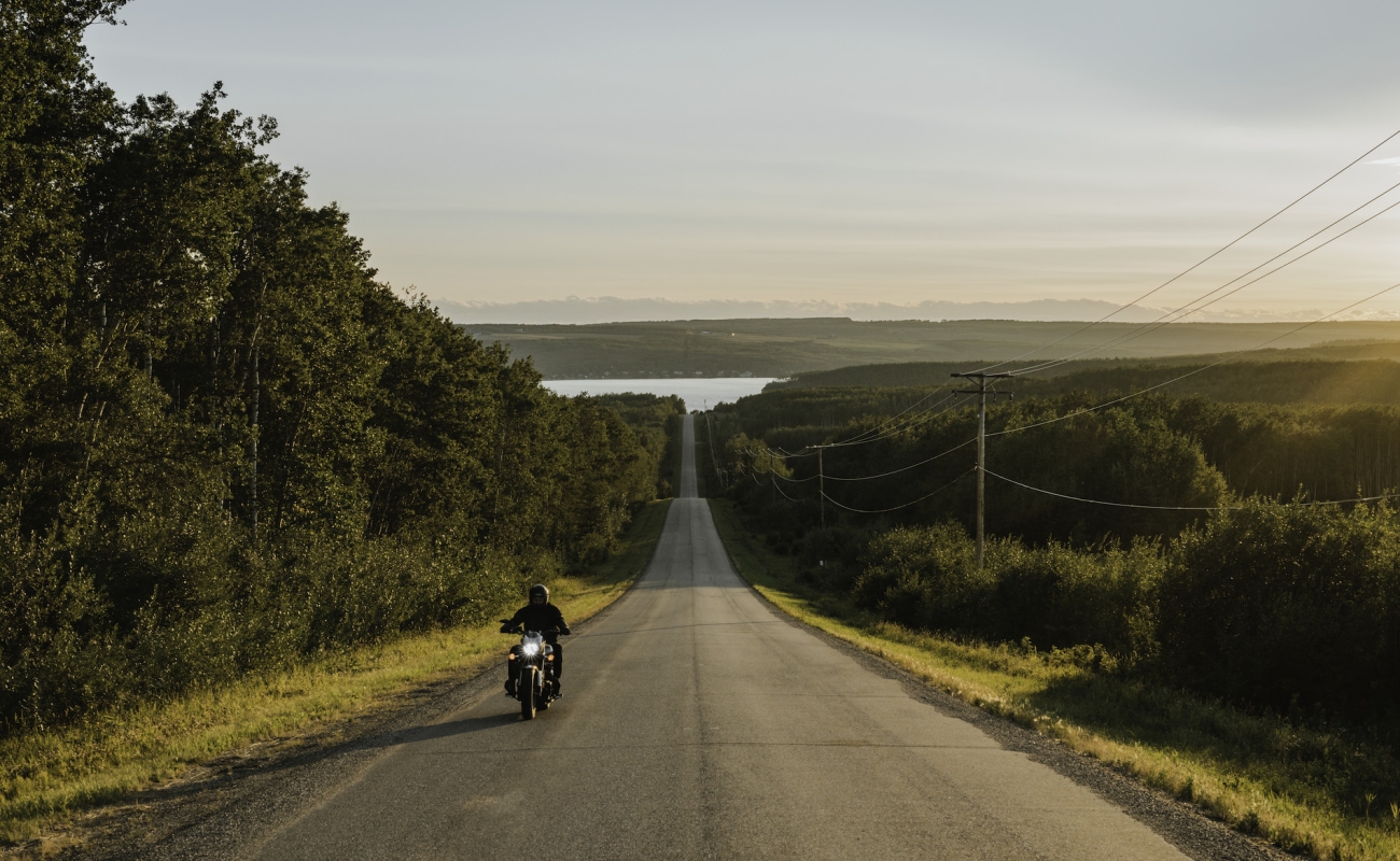 A motorocycle drives along a country road lined by foliage near Fort St. John.