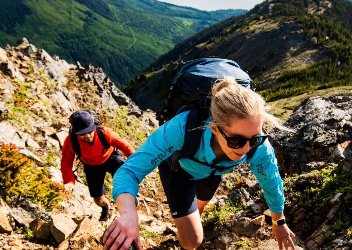 Two people scramble up a steep embankment of moss covered rock with an expansive mountain range and lake in the background.