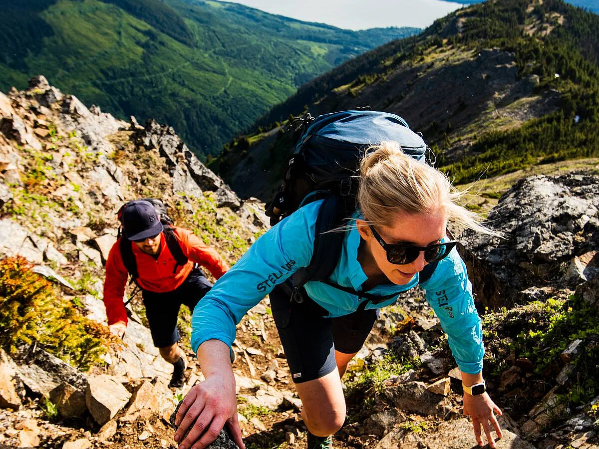 Two people scramble up a steep embankment of moss covered rock with an expansive mountain range and lake in the background.