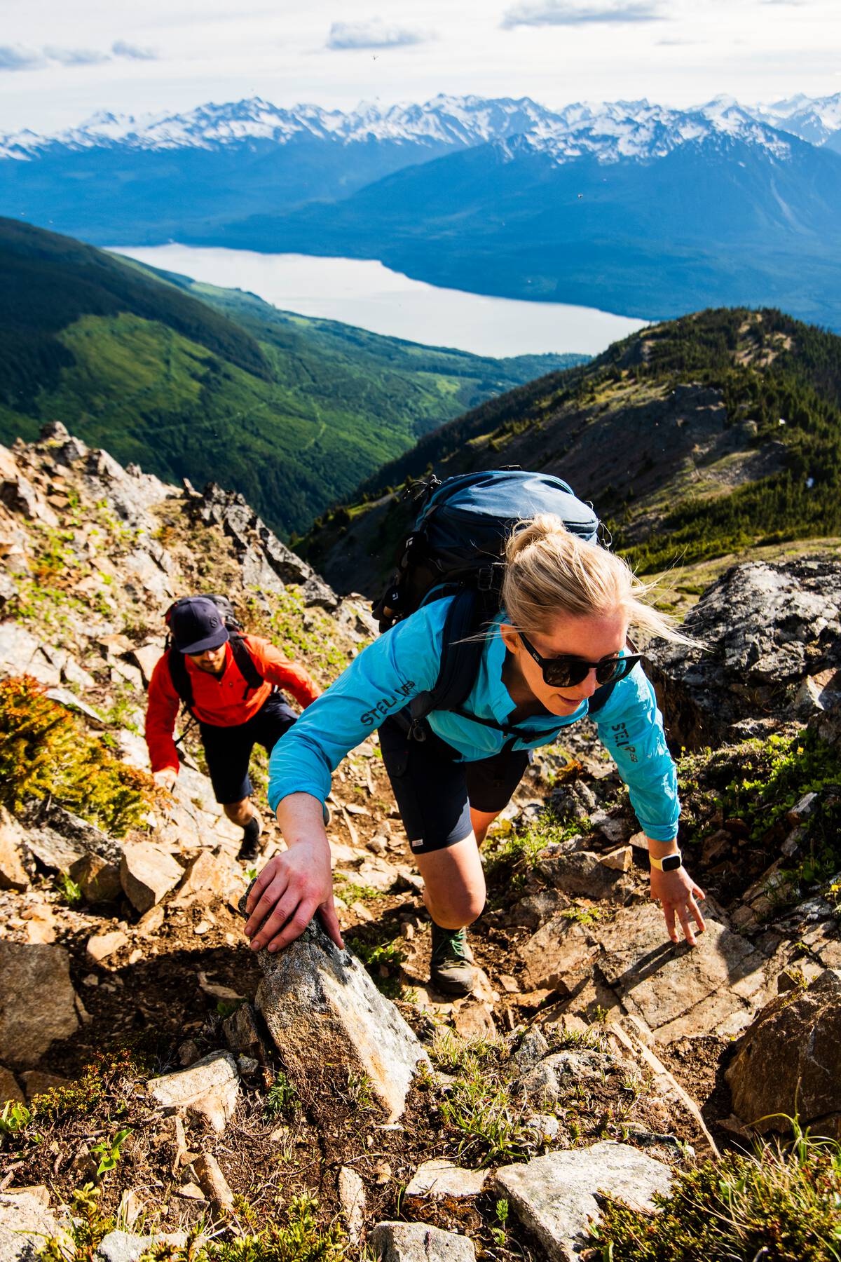 Two people scramble up a steep embankment of moss covered rock with an expansive mountain range and lake in the background.