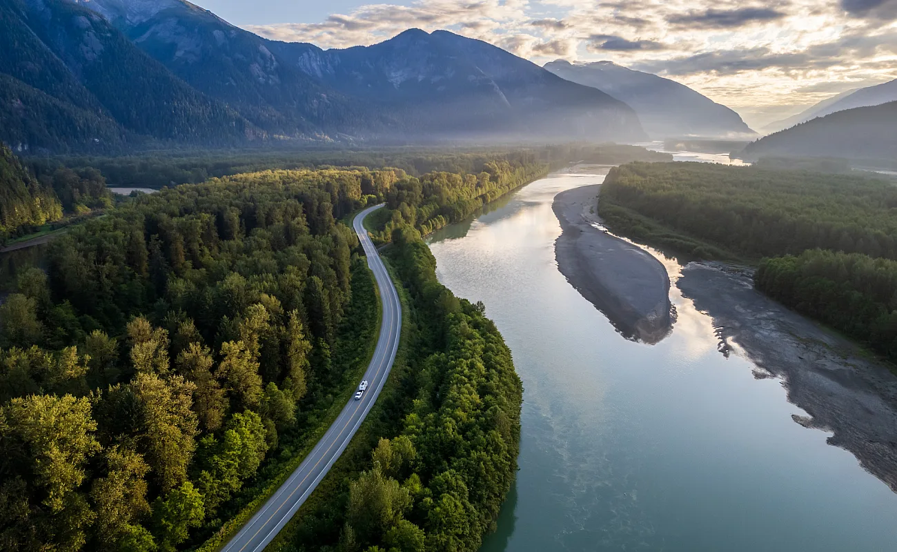 An RV winds its way along a highway, surrounded by dense forest, tall mountains and a calm river.