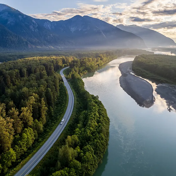 An RV winds its way along a highway, surrounded by dense forest, tall mountains and a calm river.