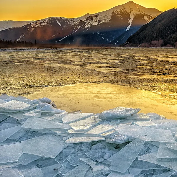Dünne Eisplatten türmen sich bei Sonnenaufgang auf der Oberfläche eines Sees auf, Berge im Hintergrund.