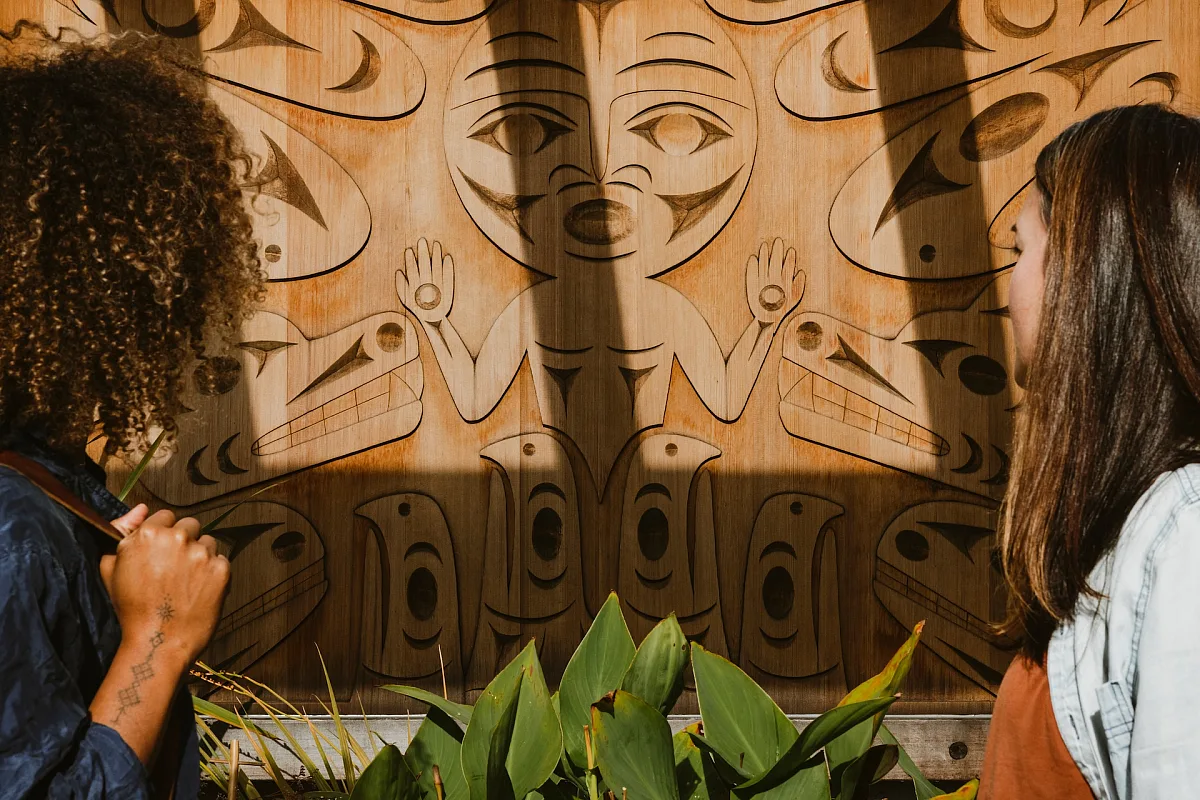 Zwei Personen betrachten die Außenwand der Gibsons Public Art Gallery. Die Schnitzerei heißt „Welh Áynexw tl’a Swa7ámchet“ oder „Spirit of Our Ancestors“. Künstler*in: Sinàmkin (Jody Broomfield) von der Squamish Nation.