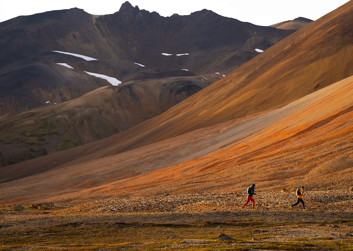 Two people wearing bright hiking attire walk along a trial beside an orange, mineral-streaked mountain.