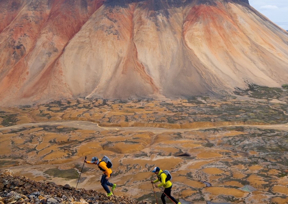 Two trail runners dash across a lunar-like landscape of orange and brown mineral mountains and grey rock.