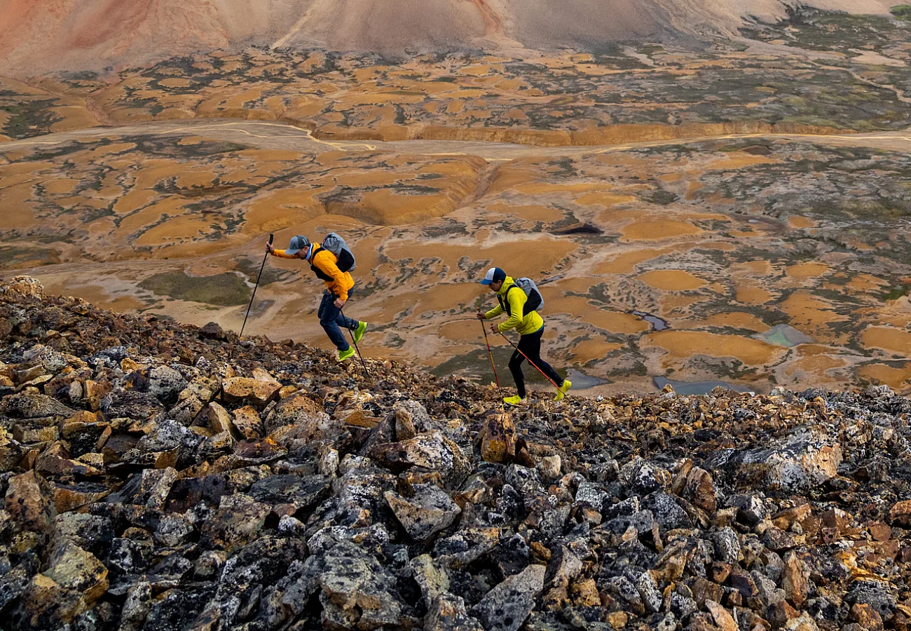 Two trail runners dash across a lunar-like landscape of orange and brown mineral mountains and grey rock.