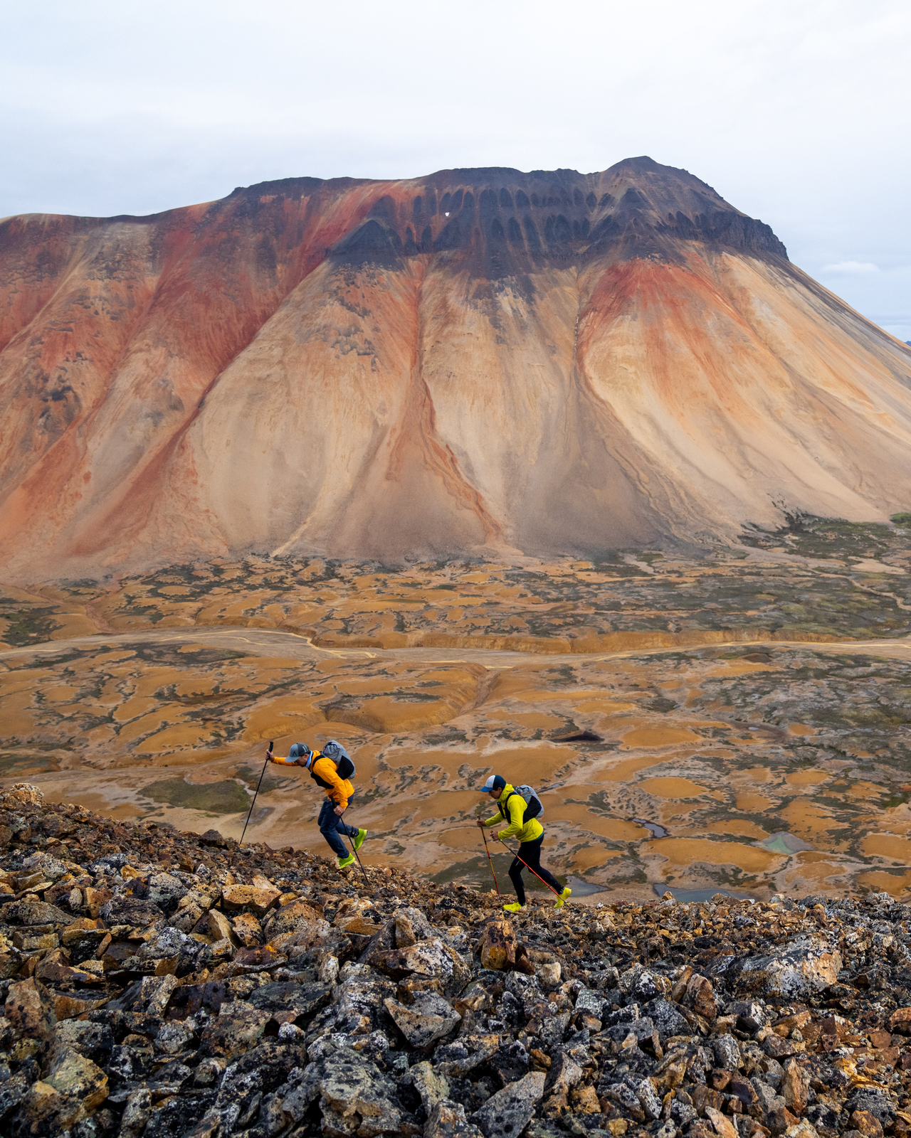 Two trail runners dash across a lunar-like landscape of orange and brown mineral mountains and grey rock.