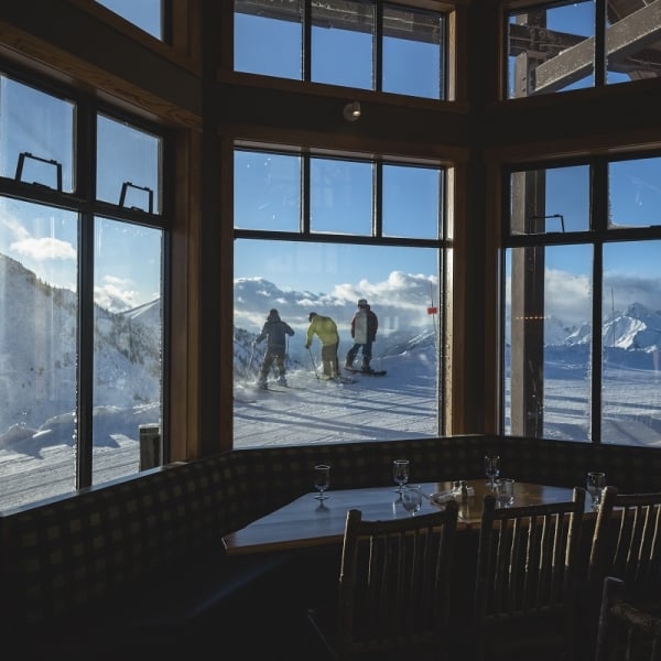 A group of three skiers head out as seen through a window in a restaurant. The restaurant is empty and dark, the ski hill is brightly lit on a blue sky day.