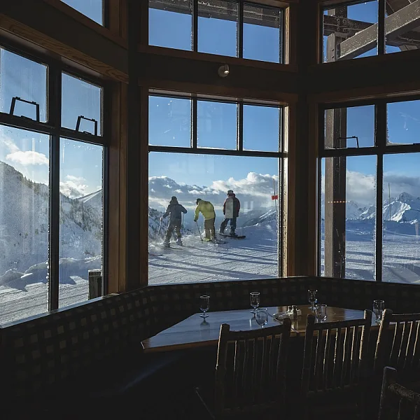 A group of three skiers head out as seen through a window in a restaurant. The restaurant is empty and dark, the ski hill is brightly lit on a blue sky day.