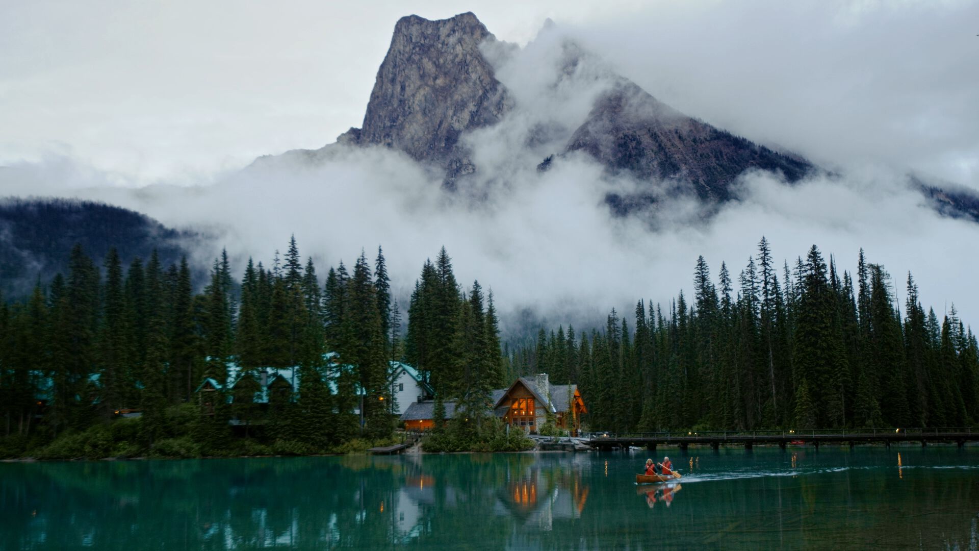 Wide shot of two people canoeing on calm lake in early evening, mountain and lights glowing in lodge in background