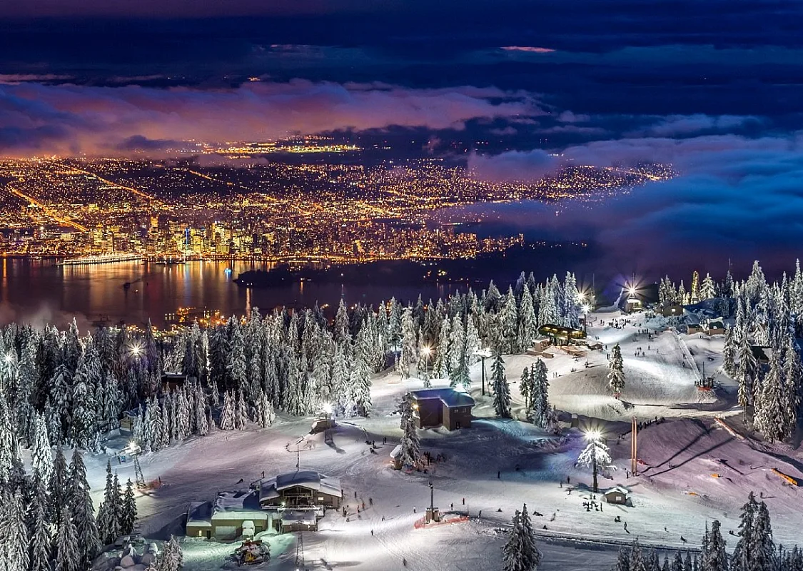 View of Vancouver from atop Grouse Mountain at night time. The resort is lit up with night time ski lights. The city is brightly lit with yellow lights below.