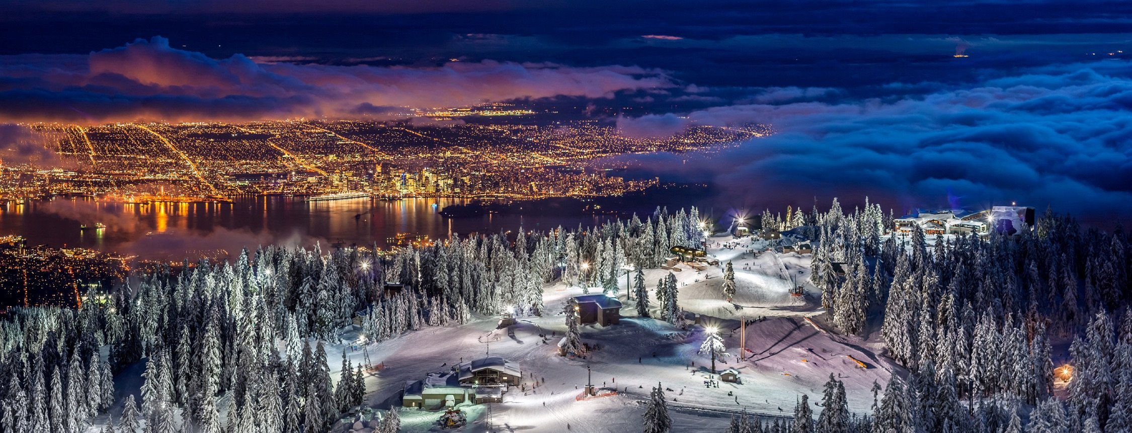 View of Vancouver from atop Grouse Mountain at night time. The resort is lit up with night time ski lights. The city is brightly lit with yellow lights below.